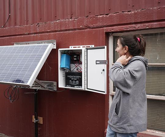 young woman with brown hair gesturing toward solar panel